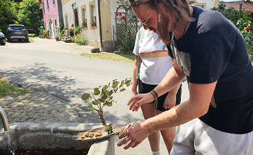 Mann setzt selbstgebautes Boot aus Naturmaterialien ins Wasser eines Brunnens.
